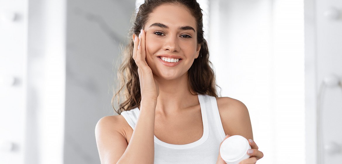 Junge Frau zeigt eine Creme, die sie sich aufs Gesicht aufträgt.
